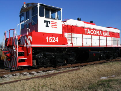 A red and white locomotive numbered 1524, labeled "TACOMA RAIL," positioned on railway tracks with grass and gravel nearby under a blue sky.