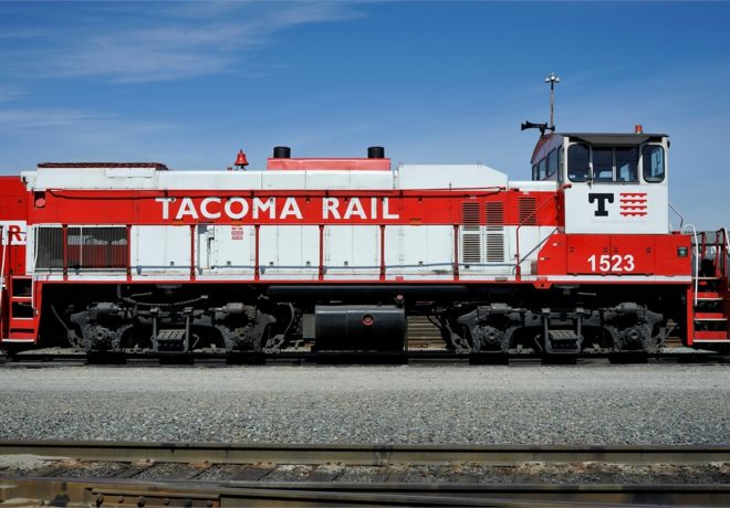 A red and white locomotive labeled Tacoma Rail 1523 on metal tracks against a blue sky.