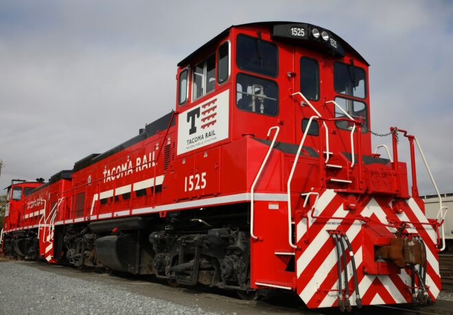 A red Tacoma Rail locomotive numbered 1525, featuring white accents and bold diagonal stripes, sits on gravel tracks under a cloudy sky.