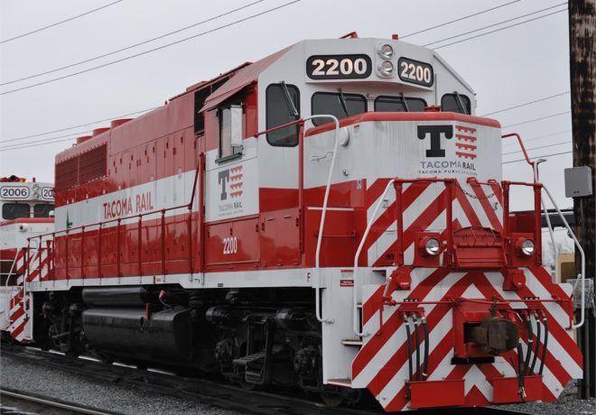Red and white diesel locomotive numbered 2200 from Tacoma Rail, featuring bold diagonal stripes and a visible logo, set in an industrial rail yard.
