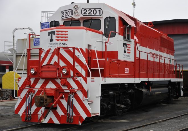 Red and white Tacoma Rail locomotive number 2201 with chevron pattern on the front, set against an industrial background.