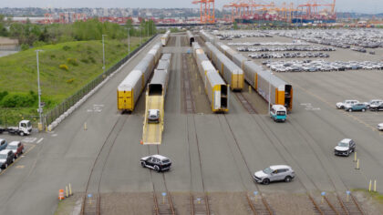 Railway yard with freight train cars and a loading ramp for vehicles; a silver car is on the ramp, nearby is a parking lot with several white vehicles and a port in the background.