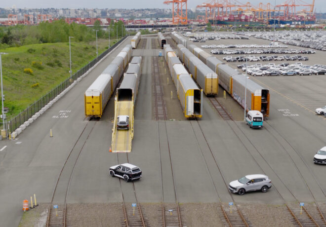 Railway yard with freight train cars and a loading ramp for vehicles; a silver car is on the ramp, nearby is a parking lot with several white vehicles and a port in the background.