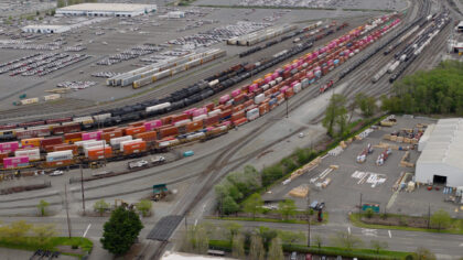 Aerial view of a freight yard featuring rows of colorful shipping containers, black tank cars, and a large parking lot filled with vehicles.