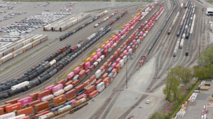 Aerial view of a freight yard featuring rows of colorful shipping containers, black tank cars, and a large parking lot filled with vehicles.