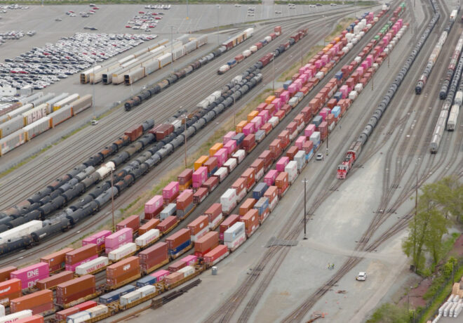 Aerial view of a freight yard featuring rows of colorful shipping containers, black tank cars, and a large parking lot filled with vehicles.