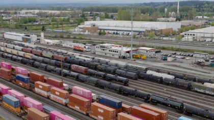Tacoma Rail freight yard filled with colorful shipping containers and black tank cars, featuring a building with a red roof and various parked vehicles in the background.