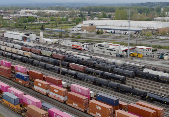 Tacoma Rail freight yard filled with colorful shipping containers and black tank cars, featuring a building with a red roof and various parked vehicles in the background.