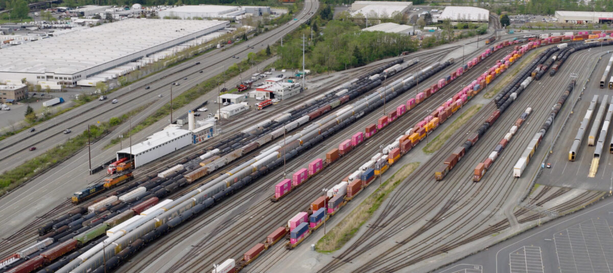 Aerial view of the Tacoma rail yard featuring numerous colorful train cars along railway tracks, with industrial buildings and a highway in the background.