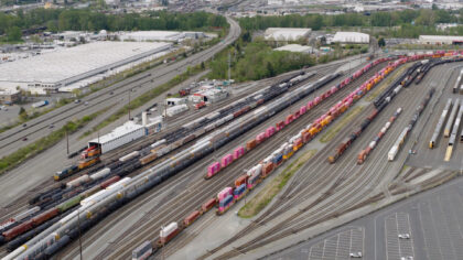 Aerial view of the Tacoma rail yard featuring numerous colorful train cars along railway tracks, with industrial buildings and a highway in the background.