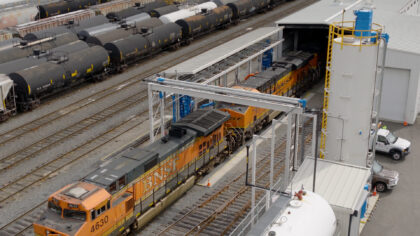 A railway yard featuring two BNSF locomotives, multiple black tank cars, and an industrial structure with a storage tank in the background.