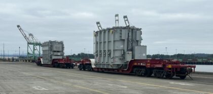 Two large electrical transformers loaded on heavy-duty railcars at a port terminal, with cranes in the background.