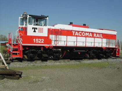 Red locomotive with "TACOMA RAIL" branding and model number "1522," positioned on a gravel track under a clear blue sky.