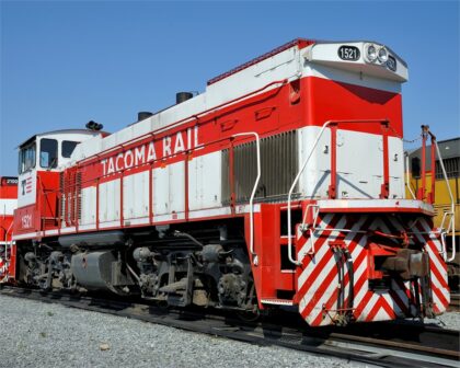 A red and white locomotive labeled Tacoma Rail 1521 on metal tracks against a blue sky.