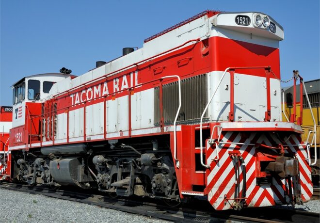 A red and white locomotive labeled Tacoma Rail 1521 on metal tracks against a blue sky.