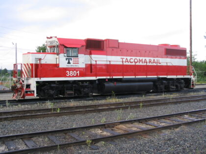 Red and white Tacoma Rail locomotive 3801 on train tracks surrounded by gravel and sparse vegetation.