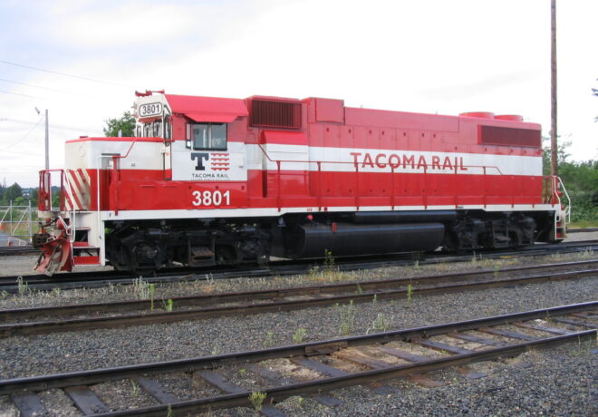 Red and white Tacoma Rail locomotive 3801 on train tracks surrounded by gravel and sparse vegetation.
