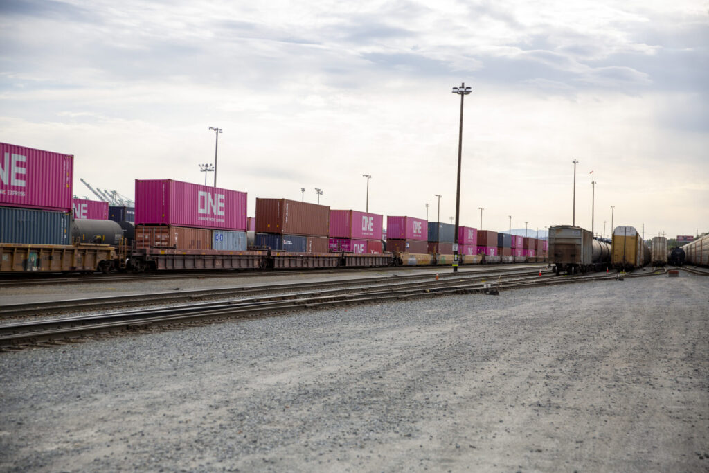 The Tacoma Rail, rail yard with stacked freight containers in various colors, notably pink with "ONE" printed in white, alongside train cars on tracks under a cloudy sky.