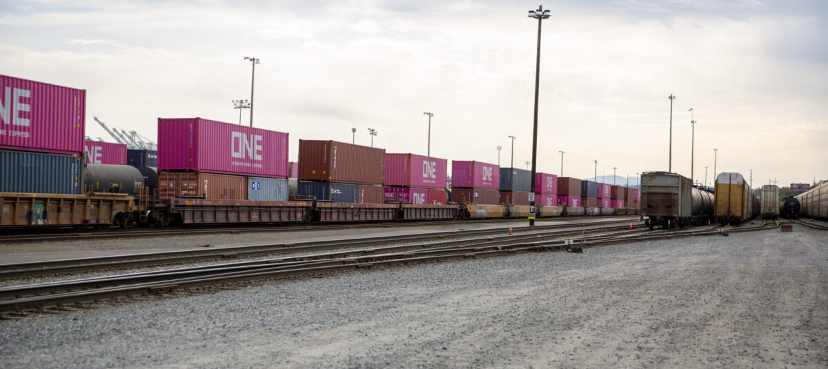The Tacoma Rail, rail yard with stacked freight containers in various colors, notably pink with "ONE" printed in white, alongside train cars on tracks under a cloudy sky.