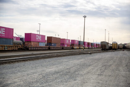 The Tacoma Rail, rail yard with stacked freight containers in various colors, notably pink with "ONE" printed in white, alongside train cars on tracks under a cloudy sky.