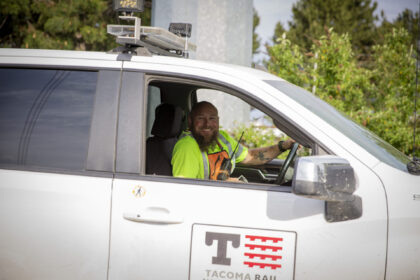 A smiling man in a bright yellow shirt and orange safety vest sits in the driver’s seat of a white vehicle, holding a radio. The vehicle is marked with a Tacoma Rail logo.