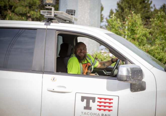 A smiling man in a bright yellow shirt and orange safety vest sits in the driver’s seat of a white vehicle, holding a radio. The vehicle is marked with a Tacoma Rail logo.