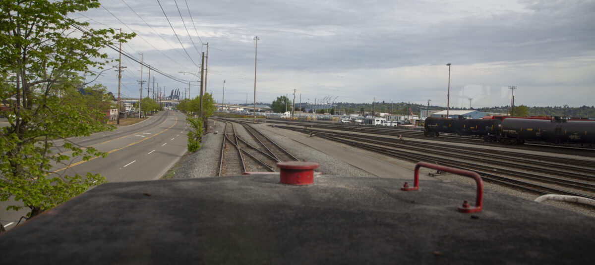 View from a train's perspective overlooking railway tracks alongside a road, with a tree and industrial structures in the background.