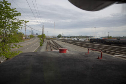 View from a train's perspective overlooking railway tracks alongside a road, with a tree and industrial structures in the background.