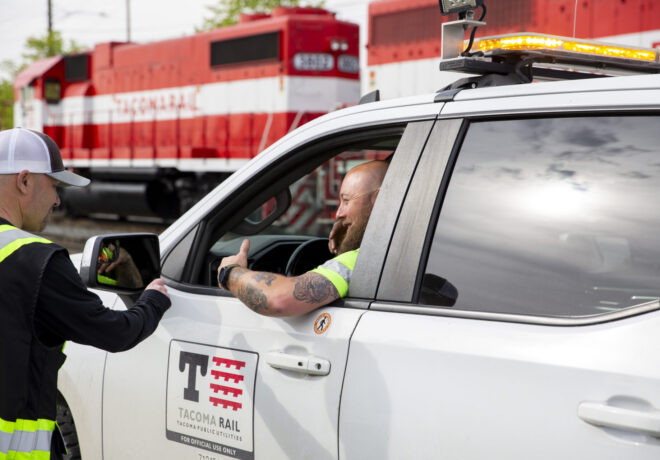A white Tacoma Rail vehicle is stopped at a railroad crossing. A smiling man in the driver's seat gives a thumbs-up, while another man in a reflective vest leans into the window, engaging with him. Red and white locomotives are visible in the background.