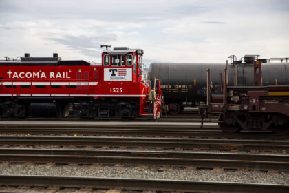 A red Tacoma Rail locomotive numbered 1525 is positioned on train tracks, with a worker in a safety vest nearby and a gray oil tank car in the background.