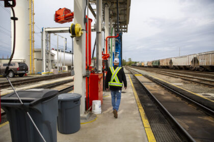 A worker in a safety vest walks along the Tacoma rail yard pathway next to freight train cars and utility equipment.