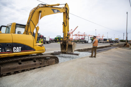 A yellow excavator at a construction site with a man in a safety vest nearby, surrounded by trucks and agricultural machinery under an overcast sky.
