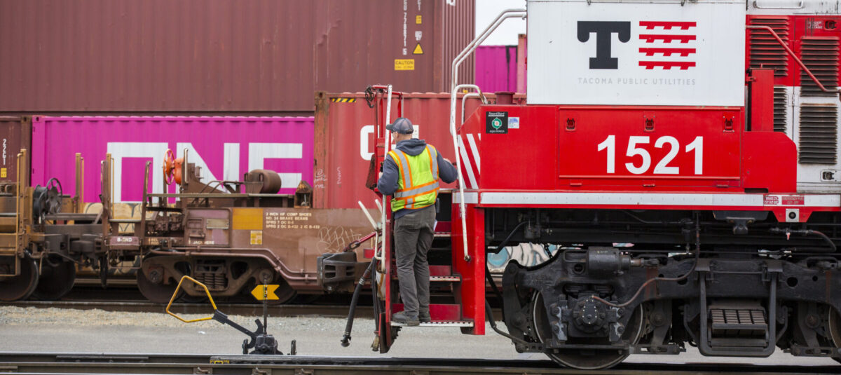 A Tacoma Rail employee in a safety vest standing on the stairs of a red and white locomotive labeled "1521" beside shipping containers, including one marked "ONE."