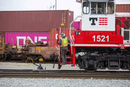 A Tacoma Rail employee in a safety vest standing on the stairs of a red and white locomotive labeled "1521" beside shipping containers, including one marked "ONE."
