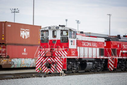 A red Tacoma Rail locomotive labeled "1521" is shown in a rail yard, surrounded by shipping containers and gravel.