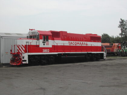 Red and white diesel locomotive number 3802 from TACOMARAIL parked beside a building in an industrial area.