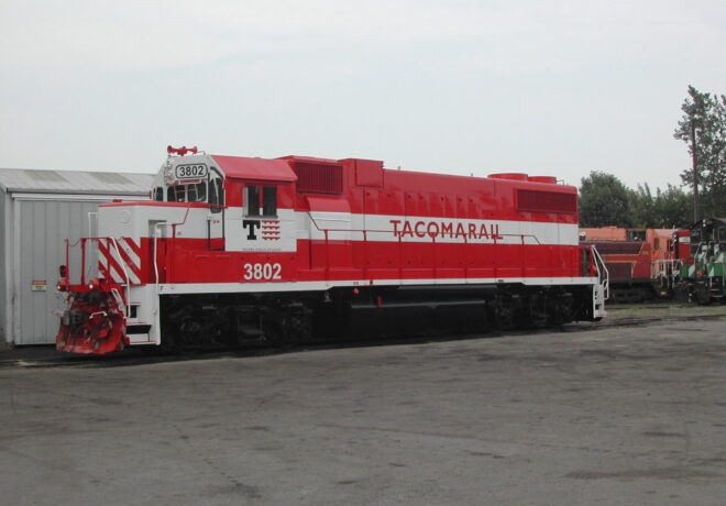 Red and white diesel locomotive number 3802 from TACOMARAIL parked beside a building in an industrial area.